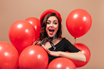Naughty green-eyed lady with red lips shouts joyfully, looks into camera and poses on beige background with balloons