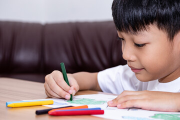 A little Asian boy is happily drawing a house with crayons is a beautiful imagination painting. A little child is engaged in creativity, the child does his homework.