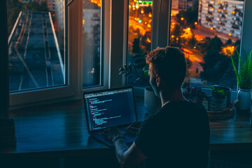 A young man programmer coding on a laptop in the dark with a view of the lights of the night city, color lighting in the room. Coding and hacking concept