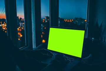 A laptop with an empty green screen on a wooden table at night with a view of the city with colored lights. A place for text, the concept of programming and development