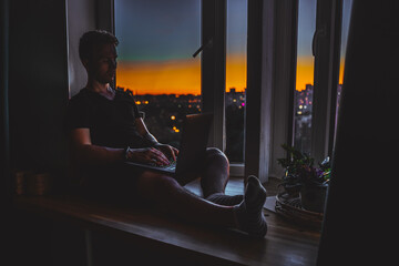 A young programmer is coding on a laptop in the dark sitting on a window with a view of the lights of the night city, colored lighting in the room, home decor