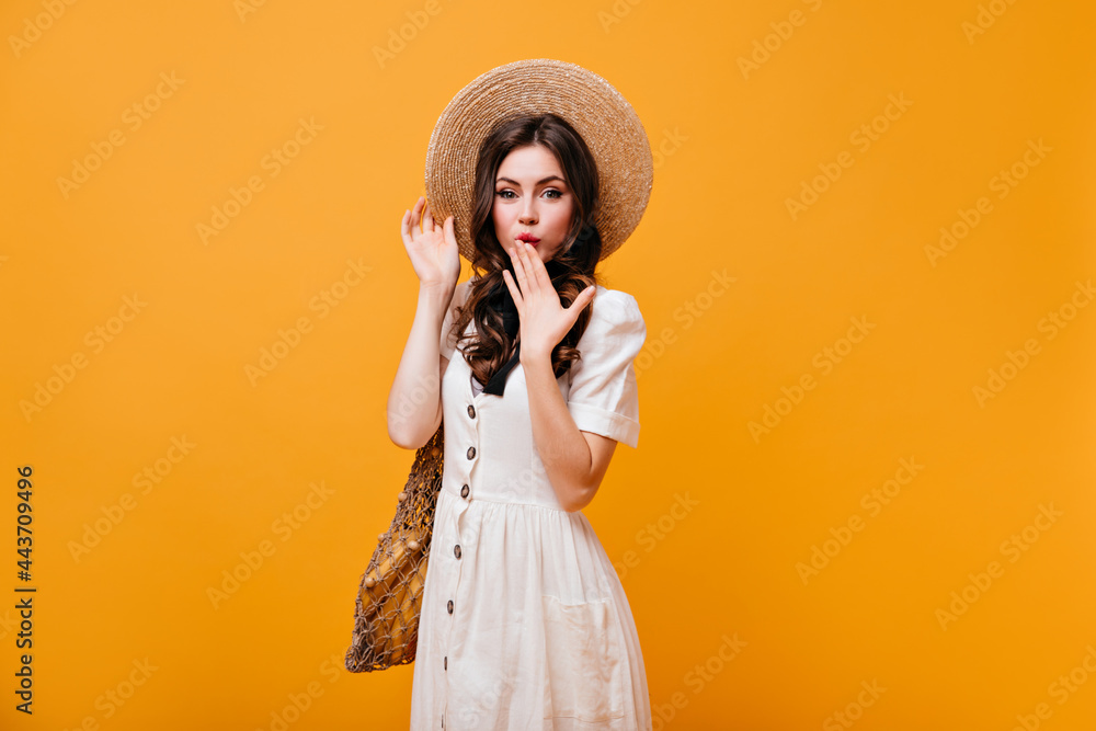 Wall mural Green-eyed lady covers her mouth with her hand. Woman in straw hat and white sundress holds shopping bag on orange background