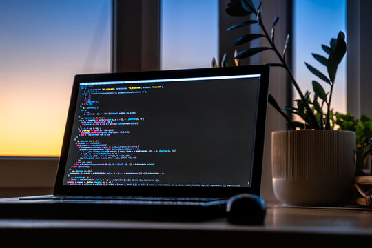 The code is on a laptop on a wooden table in front of the window  in the dark with a view of the lights of the night city, color lighting in the room, home decor	
