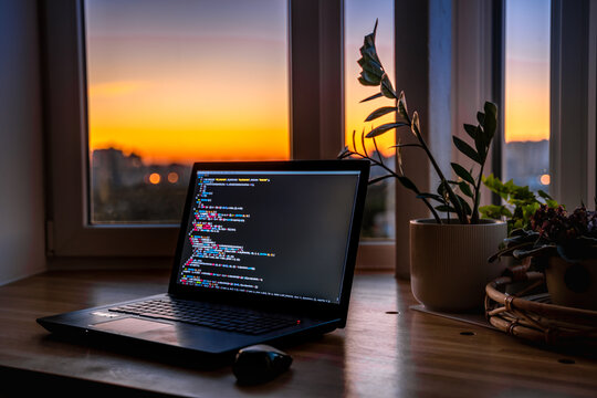The code is on a laptop on a wooden table in front of the window  in the dark with a view of the lights of the night city, color lighting in the room, home decor	