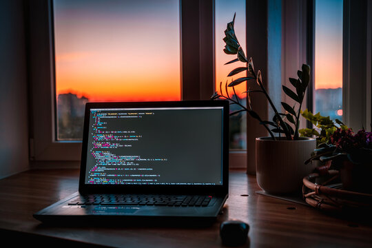 The code is on a laptop on a wooden table in front of the window  in the dark with a view of the lights of the night city, color lighting in the room, home decor	