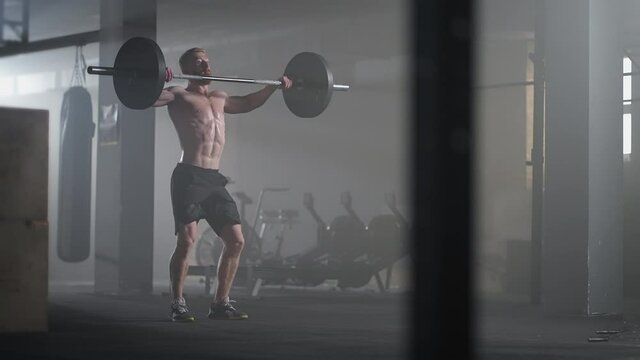 Slow motion: Brutal athlete lifts the bar above himself, performing a jerk, a spinning push. A man is engaged in weightlifting on a dark background, portrait. Concept strength, power, playing sports.