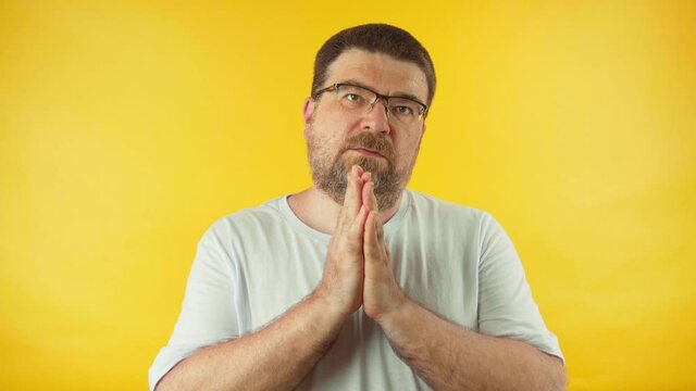Caucasian middle aged man in white t-shirt praying over yellow background. Man begging someone satisfy his desires, help with.