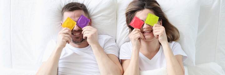 Man and woman are holding colorful condoms while lying on bed
