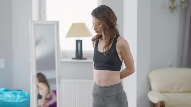 Beautiful slim Caucasian woman measuring waist making dissatisfied facial expression. Portrait of sad millennial disappointed with weight loss result standing indoors thinking