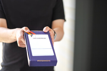 Messenger carry box package with first class label at front door, depth of field. Man in black clothes deliver priority packet at customer’s house, blurred background. Express delivery parcel concept
