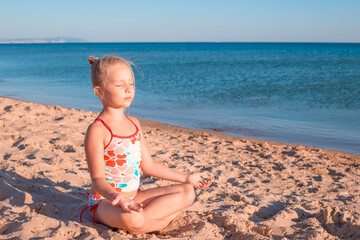 A little girl sits on the sand in a lotus position and meditates on the seashore. Yoga classes in nature