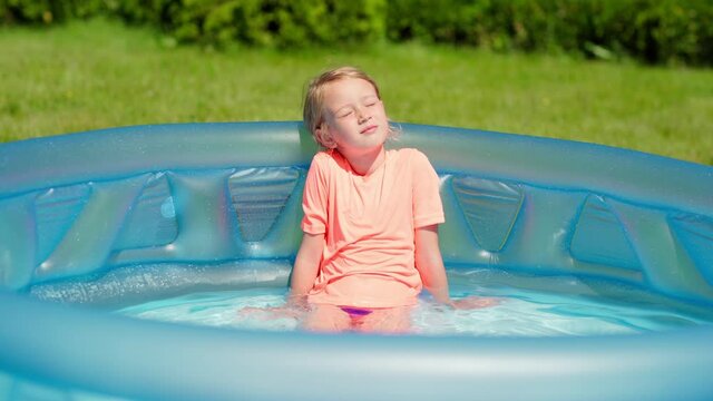 Tilt up of cute little girl relaxing and sunbathing with closed eyes in inflatable swimming pool on sunny summer day. Child splashing water with her legs