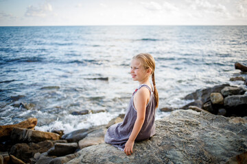 A blonde girl in a sundress sits on a stone on the seashore.