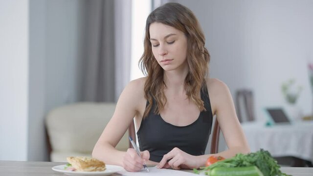 Concentrated slim motivated young woman writing calories looking at burger on plate. Portrait of Caucasian millennial controlling weight taking care of health. Confidence and self-control