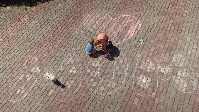 Tilt down top view of little girl and toddler boy drawing word MOM and heart on pavement near house on mothers day or mum's birthday. Chihuahua dog walking nearby. Kids preparing surprise for mother