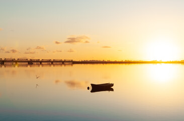 Sunrise across bay with golden glow and moored dinghy and railway bridge in distance.