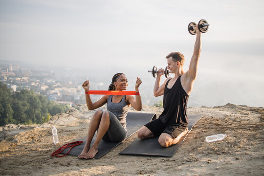 African Woman In Sport Clothes Training Arms Using Rubber Band While Strong Caucasian Man Workout With Heavy Dumbbells. Active Couple Exercising Outdoors.