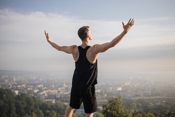 Fototapeta na wymiar Back close up view of Caucasian male athlete relaxing during outdoors workout. Muscular young man in sport clothes standing with hands outstretched on high hill and enjoying cityscape view.