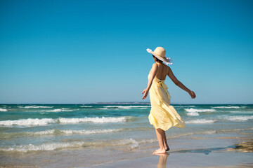 Woman with straw hat and yellow dress on the beach and arms streched.