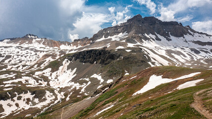 View of mountain peaks in Yankee Boy Basin near Ouray, Colorado. The mountain range has snow on it with jagged rocks, a blue cloudy sky, and green grass.