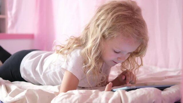 Caucasian curious curly cute preschool kid girl using digital tablet technology device lying on bed alone. Small child hold pad surfing internet play game at home. Children tech addiction 
