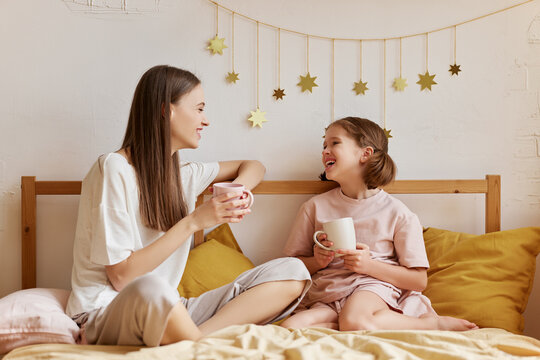 Two Girls, Cheerful Older And Younger Sister Drinking Tea And Talking While Spending Time Together In Bedroom