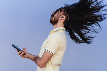 young, long-haired man using a cell phone with fast internet, using wireless headphones, dazzled by...