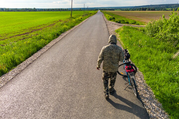 A cyclist in camouflage from the back next to a bicycle on the background of an asphalt road going...