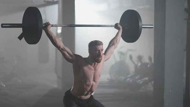 Slow motion: Brutal athlete lifts the bar above himself, performing a jerk, a spinning push. A man is engaged in weightlifting on a dark background, portrait. Concept strength, power, playing sports.