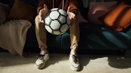 Young man holding soccer ball and sitting on sofa in living room, sport portrait at home, guy football fan twirling ball on his finger, teen is ready for watching professional game on television. 
