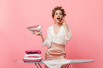 Lady talking on phone and opened her mouth in surprise. Housewife ironing clothes on pink background