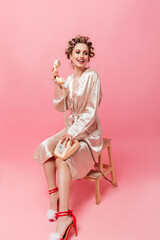 Fototapeta premium Charming girl in pink robe smiles, sits on chair and holds phone on isolated background.
