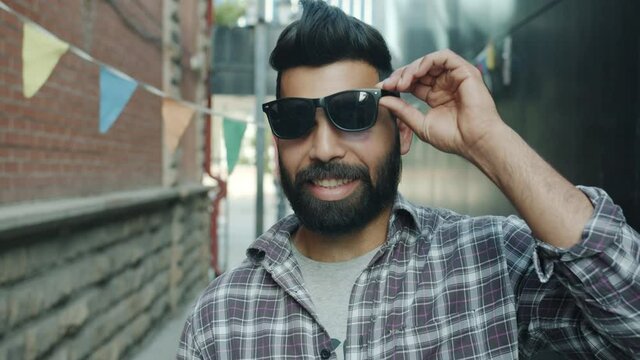 Portrait of stylish Arab guy raising sunglasses smiling and winking outdoors in city street. Facial expressions and modern young people concept.