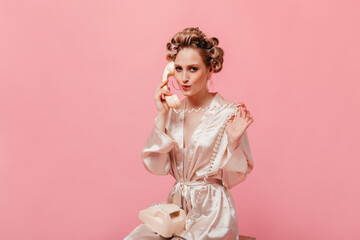 Beautiful woman in silk dressing gown coquettishly talking on phone on pink background