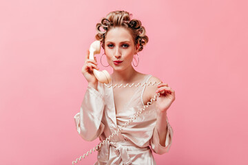 Flirtatious woman with hair curlers bites her lip and talks on landline phone on pink background.