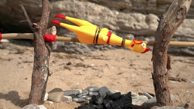Funny scene of cooking by roasting on spit toy chicken on artificial bonfire on beach against blurred rocks ON sunny day closeup