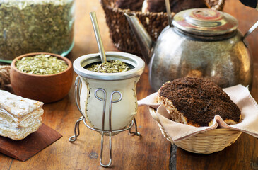 Mate drink over a wooden table with old teapot, pastries and bowl and jar with yerba mate sticked dried leaves