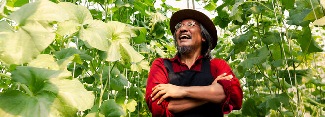Senior middle aged male farmer having arms crossed with happy teethy smile wearing a straw hat in red farming uniform inside farm garden in summer - 443741457