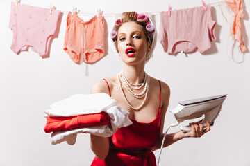 Blond woman in pearl necklace and hair curlers poses with iron and linen
