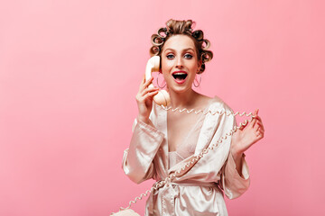 Portrait of blue-eyed woman with hair curlers looking at camera, talking on phone and coquettishly twirling wire on her finger.