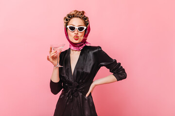 Fototapeta premium Lady in sunglasses posing on pink background, holding martini glass and eating olive