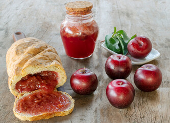Sweet bread stuffed with red Plum jam over a wood board over a table with fresh red Plums and a jar with Plums confiture