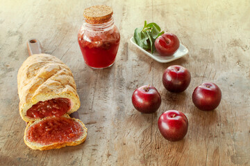 Sweet bread stuffed with red Plum jam over a wood board over a table with fresh red Plums and a jar with Plums confiture