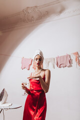 Gorgeous woman in red outfit is holding martini glass and posing against backdrop of drying clothes and iron