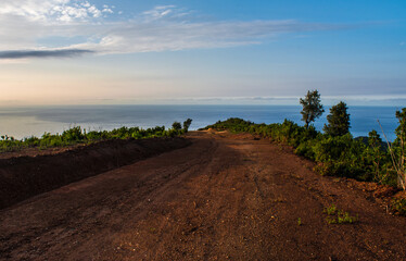 A mountainous dirt road overlooking the sea, a bumpy road with red soil amidst forest trees and blue sky facing the sea with sunset, wandering among the beautiful green nature.
