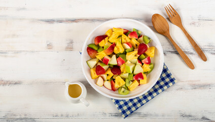 Mixed fruits salad including strawberry, kiwi, apple, and pineapple in white dish place on sackcloth on wooded table.  Utensil spoon and fork and oil salad dressing cup beside