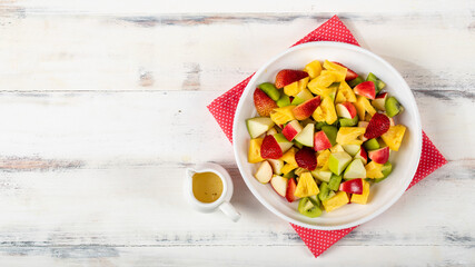 Mixed fruits salad including strawberry, kiwi, apple, and pineapple in white dish place on sackcloth on wooded table.  Utensil spoon and fork and oil salad dressing cup beside