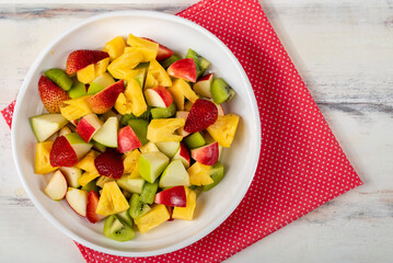 Mixed fruits salad including strawberry, kiwi, apple, and pineapple in white dish place on sackcloth on wooded table.  Utensil spoon and fork and oil salad dressing cup beside