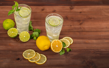 Two refreshing glasses of cold fruit juice, lime and lemon with ice, decorating the glasses with mint leaf. Fresh lime and lemon both cut and slice are on wooden table behind red brick wall