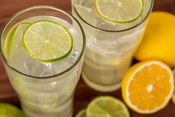 A close up picture of two refreshing glasses of cold fruit juice with straw, ice lime and lemon juice, decorating with fresh lime and lemon on wooden table. Concept for refreshing